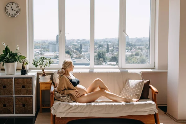 孕妇穿着舒适的羊毛衫 坐在家里靠窗的柔软光线下的沙发上 — 图库照片