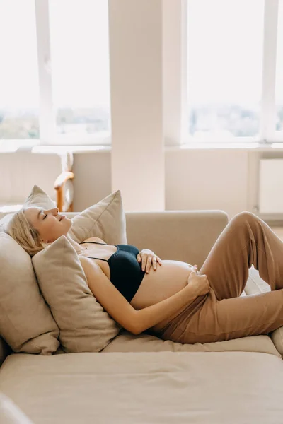 妊娠中の女性は居心地の良い家庭用の服を着て 軽い室内のソファに横になり 腹を手で握って — ストック写真