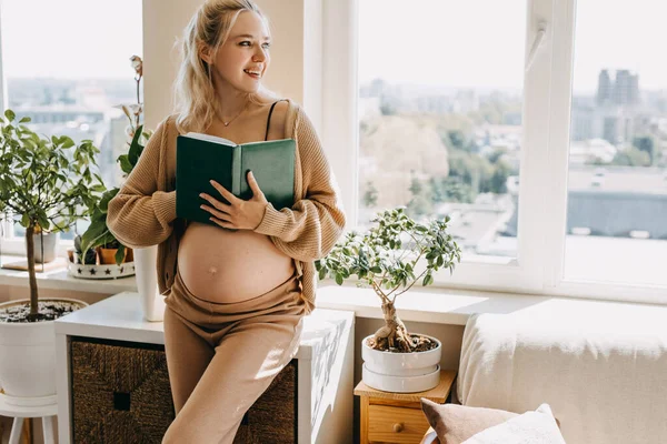 妊娠中の女性は居心地の良い家庭用の服を着て本を持ち笑顔で — ストック写真