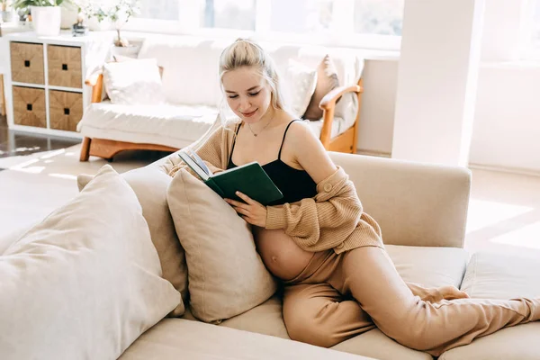 居心地の良い家庭用の服を着て家のソファに座って本を読んでいる妊婦 — ストック写真