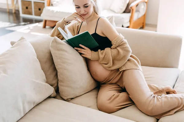 ブロンド妊婦を着て家で居心地の良いソファに座って本を読む — ストック写真