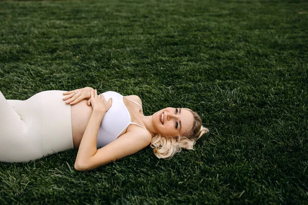 ブロンド妊娠中の女性スポーツウェアを着て緑の芝生の上に横になり リラックスし 腹に手を握って — ストック写真