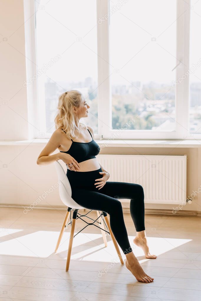 Blonde pregnant woman wearing black top and leggins, sitting on a chair at home, looking through the window.