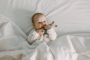 Bebek beyaz bir battaniyeyle yatakta uzanıyor, oyuncak tutuyor, parmaklarını emiyor..