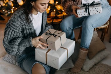 Sarılıp sarmalanmış hediyeler taşıyan genç esmer kadın, Noel ağacı arka planında oturuyor..