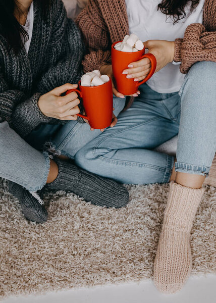 Closeup of two women wearing knitted sweaters and socks, at home, with red cups with cocoa and marshmallows.