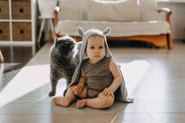 Bebek evde kedisiyle yerde oturuyor, kapüşonlu hırka giyiyor ve tavşan kulağı takıyor..