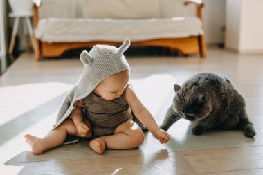 Bebek yerde oturup kediyle oynuyor, kulakları olan kapüşonlu bir hırka giyiyor..