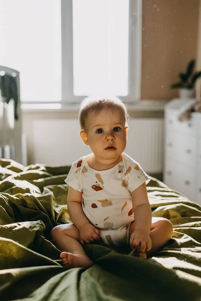 Little 9 months old baby sitting on bed at home, in sun light, looking at camera.