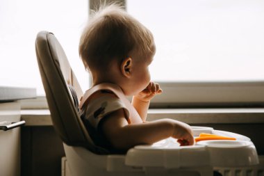 10 aylık bebek yüksek sandalyede oturuyor, pencerenin kenarında parmak yiyeceği yiyor..