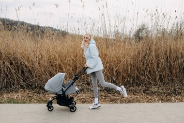 Baharda göl kenarında bebek arabasıyla yürüyen mutlu sarışın kadın.. 