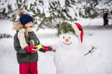 Karlı kış yürüyüşünde bir kardan adamla oynaşan mutlu sarışın kız çocuğu.
