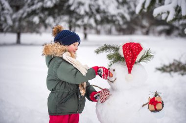 Karlı kış yürüyüşünde bir kardan adamla oynaşan mutlu sarışın kız çocuğu.