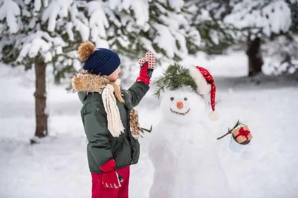 Karlı kış yürüyüşünde bir kardan adamla oynaşan mutlu sarışın kız çocuğu.