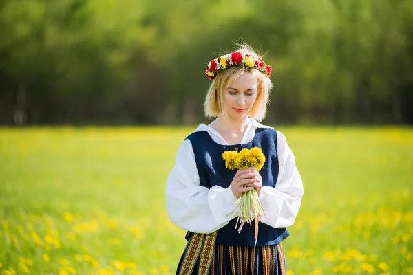 Egy fiatal nő nemzeti ruhában sárga pitypang koszorút visel a tavaszi mezőn. Tavasz — Stock Fotó