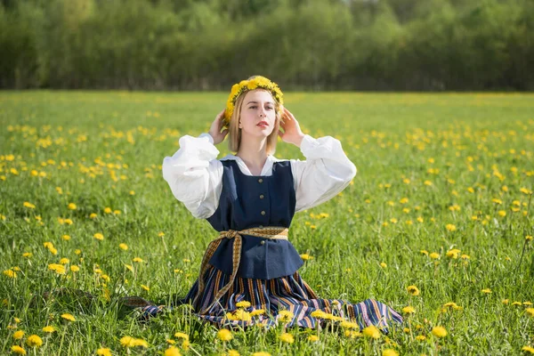 Jovem mulher em roupas nacionais vestindo coroa de dente de leão amarelo no campo de primavera. Ligo... — Fotografia de Stock