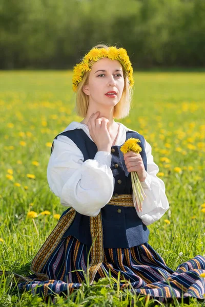 Mladá žena v národních šatech se žlutou pampeliškou na jarním poli. Ligo — Stock fotografie