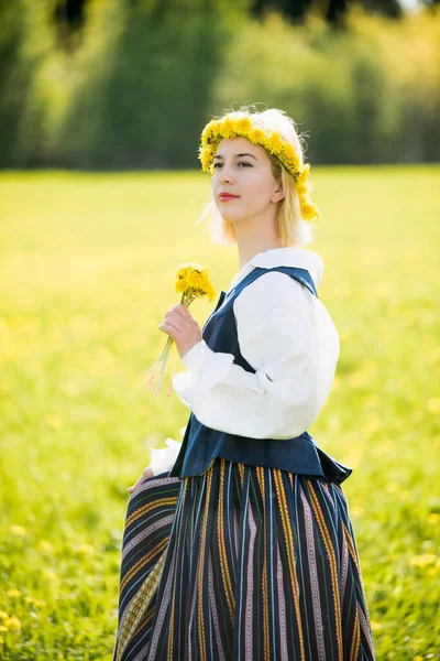 Ung kvinna i nationella kläder bär gul maskros krans på våren fält. Ligo — Stockfoto