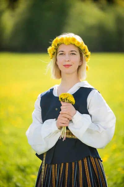 Mladá žena v národních šatech se žlutou pampeliškou na jarním poli. Ligo — Stock fotografie