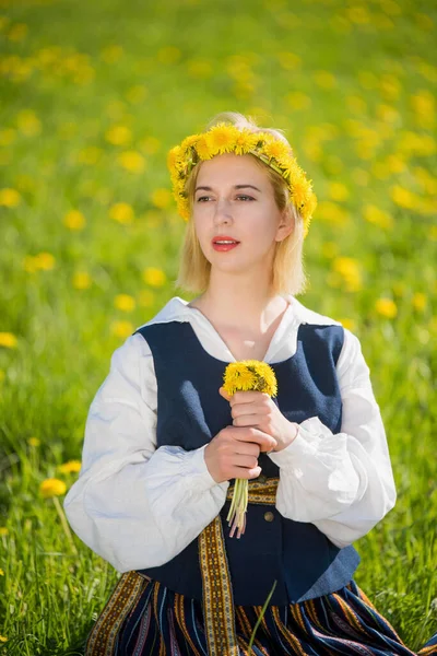 Giovane donna in abiti nazionali che indossa ghirlanda di dente di leone giallo nel campo di primavera. Ligo. — Foto Stock
