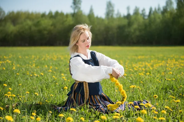 Ulusal giysili genç bir kadın bahar tarlasında sarı karahindiba çelenk giyiyor. Ligo. — Stok fotoğraf