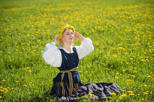 Jovem mulher em roupas nacionais vestindo coroa de dente de leão amarelo no campo de primavera. Ligo... — Fotografia de Stock