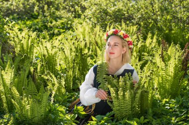Yeşil eğrelti otunun arka planında, ulusal kıyafetleri ve çelengi olan genç bir kadın. Ligo Letonya tatili
