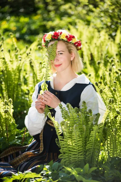 Yeşil eğrelti otunun arka planında, ulusal kıyafetleri ve çelengi olan genç bir kadın. Ligo Letonya tatili — Stok fotoğraf