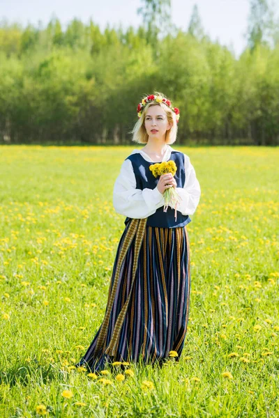 Jovem mulher em roupas nacionais vestindo coroa de dente de leão amarelo no campo de primavera. Ligo... — Fotografia de Stock