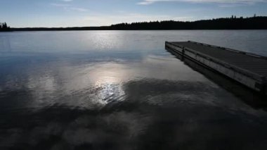 Suya yansıyan güneşle birlikte sakin büyük bir göl, sağ taraf boyunca bir rıhtım ve uzakta bir orman.