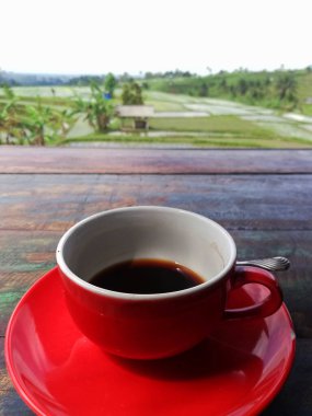 Black coffee on red color cup,placed on wooden table,with rice field background