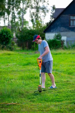 A young handsome guy in a cap with an electric lawn mower mows the grass on the lawn near the house. Small depth of field.