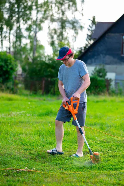 A young handsome guy in a cap with an electric lawn mower mows the grass on the lawn near the house. Small depth of field.