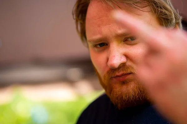 Close-up of the face of handsome man with red hair and a beard, in the foreground an out-of-focus hand with a middle finger, the background is also out of focus.