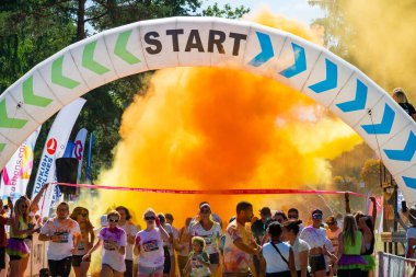 Jurmala, Kauguri, Latvia - August 14 2022: Close-up of the start of race on track with paints. Participants run in orange cloud of colored powder. Spectators behind the fence. The color run.