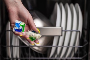 Close-up of young woman hand holding two colored capsule for the dishwasher. In the background, out of focus, is a dishwasher with clean dishes. Small depth of field.