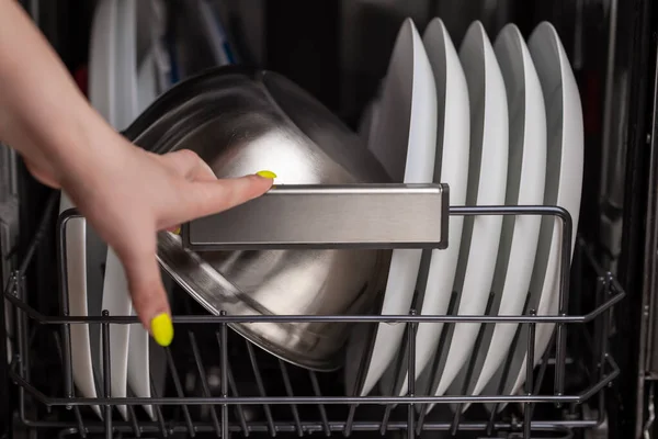 Close-up of a dishwasher basket with clean white plates and a metal bowl, a woman\'s hand pulls out clean dishes. Small depth of field.