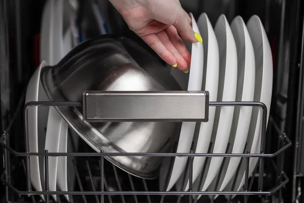 Close-up of a dishwasher basket with clean white plates and a metal bowl, a woman's hand pulls out clean dishes. Small depth of field.