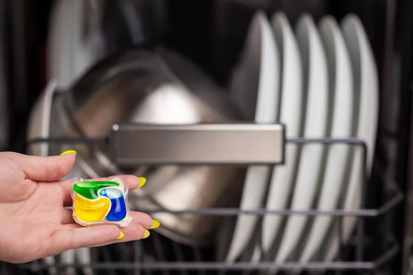 Close-up of young woman hand holding a colored capsule for the dishwasher. In the background, out of focus, is a dishwasher with clean dishes. Small depth of field.