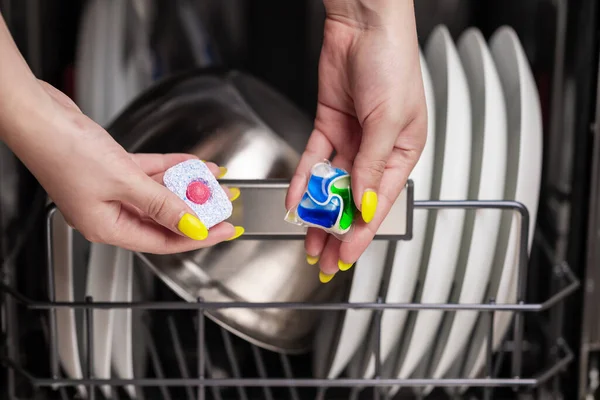 Close-up of young woman hands holding two colored capsule for the dishwasher. In the background, out of focus, is a dishwasher with clean dishes. Small depth of field.