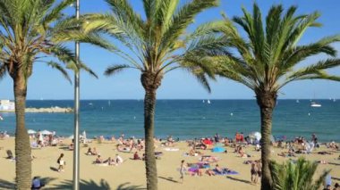 Barselona, İspanya - 29 Mayıs 2022: Birçok insanın güneşlendiği, dinlendiği ve top oynadığı Balear Denizi Sahili manzarası. Küçük tekneler suda yüzer. Palmiye ağaçlarının arasından bak. Yüksek kalite 4k görüntü
