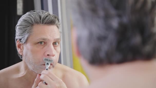 Um homem adulto de cabelos grisalhos faz a barba com uma lâmina na frente de um espelho — Vídeo de Stock