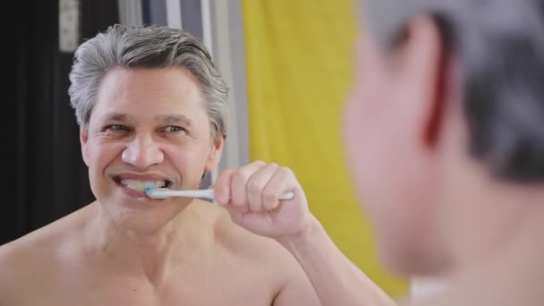 Um homem adulto de cabelos grisalhos escova os dentes com uma escova de dentes e olha no espelho — Vídeo de Stock