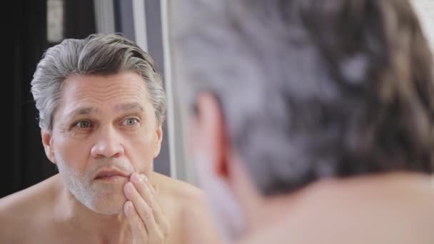 Ένας ενήλικος γκριζομάλλης άντρας βάζει κρέμα ξυρίσματος στο πρόσωπό του μπροστά από έναν καθρέφτη.. — Αρχείο Βίντεο
