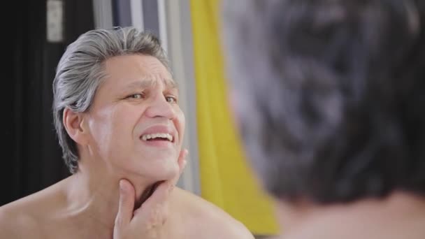Ένας ενήλικος γκριζομάλλης εξετάζει το πρόσωπό του μπροστά από έναν καθρέφτη αφού ξυριστεί και πλένει το πρόσωπό του.. — Αρχείο Βίντεο