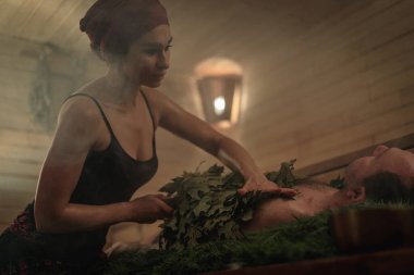 Bir kadın süpürgelerin yardımıyla bir erkek için banyo ritüeli gerçekleştiriyor. Bir adam ladin dallarına uzanır..