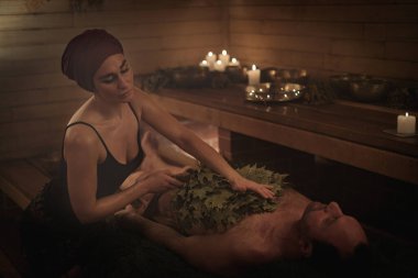 Bir kadın süpürgelerin yardımıyla bir erkek için banyo ritüeli gerçekleştiriyor. Bir adam ladin dallarına uzanır..