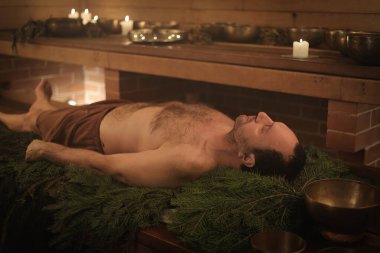 Saunadaki ladin dallarına uzanmış bir adam, Tibet ayinini gerçekleştiriyor.