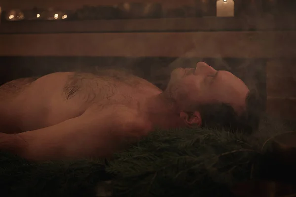 A man lies on spruce branches in a sauna, he is being performed a Tibetan ritual