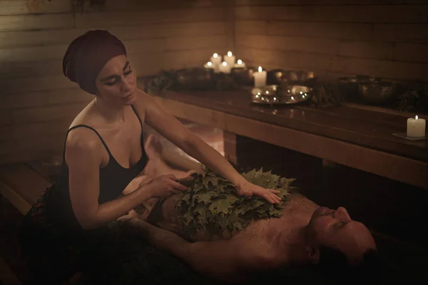 一个女人在扫帚的帮助下 为一个男人举行一个沐浴仪式 一个男人躺在云杉树枝上 免版税图库图片